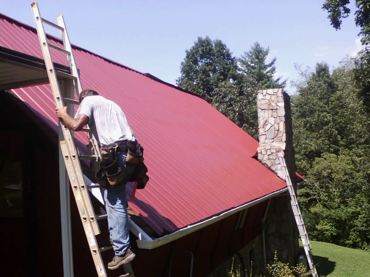 Roofer on a ladder beside a red metal roof and stone chimney — Soffit & Fascia Repair by Roofing Brothers in Big Bar